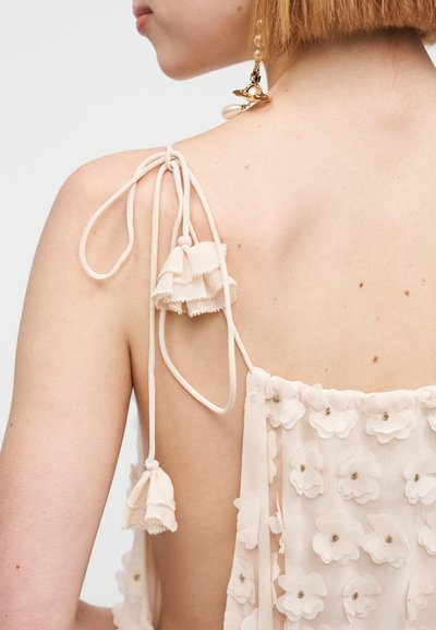 Robe beige clair avec un motif floral, détails à volants sur les bretelles et pompons décoratifs. Tissu doux et transparent.