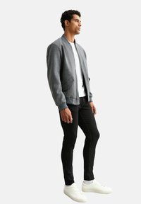 Grå bomberjacka med ribbade ärmslut, överdragande detaljer, vit t-shirt, svarta smala jeans och vita sneakers. Slät tyg och strukturerad passform.