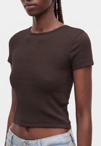 Jeune personne aux cheveux tressés foncés porte un crop top marron foncé ajusté à manches courtes et un jean bleu clair sur un fond uni.