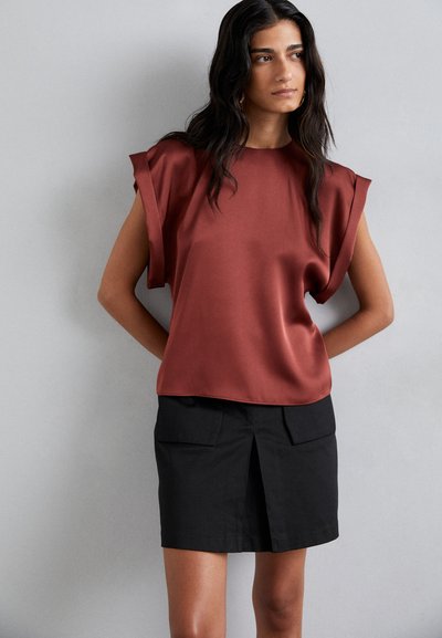 Top de satén en tono óxido con mangas anchas, combinado con una falda negra que presenta una abertura frontal y dos bolsillos delanteros. Textura suave y diseño minimalista.