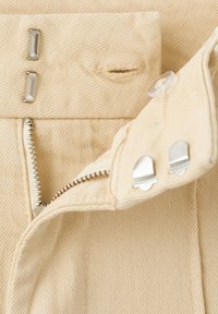 Vita di un indumento beige con cerniera metallica parzialmente aperta, occhiello, bottone trasparente in plastica e due fermagli argento a gancio e barrette.