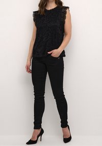Czarna koronkowa bluzka z kwiatowym wzorem i falbaniastymi brzegami, zestawiona z czarnymi obcisłymi dżinsami i spiczastymi czarnymi szpilkami. Modelka stoi z jedną ręką na biodrze.