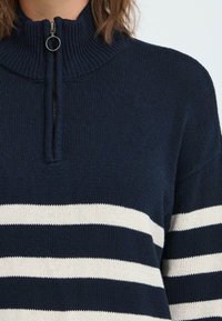 Marineblå strikkegenser med høy krage og halv glidelås. Har horisontale hvite striper og en teksturert, ribbet stoff.