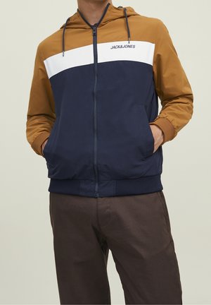 Mężczyzna ubrany w kurtkę z kapturem w kolorowe bloki z musztardowymi rękawami, białym paskiem i granatowym tułowiem, z rękami w kieszeniach, stoi na jednolitym tle.