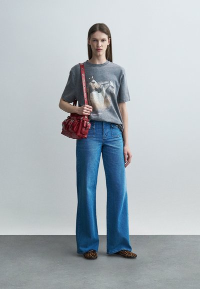 Γυναίκα που φορά γκρι μπλουζάκι με στάμπα αλόγου, μπλε παντελόνι καμπάνα, παπούτσια με λεοπάρ σχέδιο και κρατά κόκκινη τσάντα στον ώμο της.