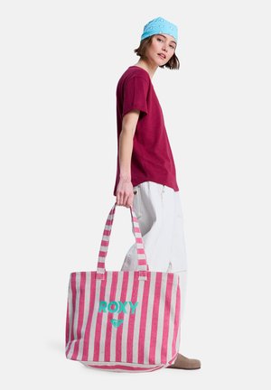 Jonge vrouw met een blauwe bandana en een bordeauxrode blouse die een grote roze-wit gestreepte draagtas vasthoudt met het "ROXY"-logo.