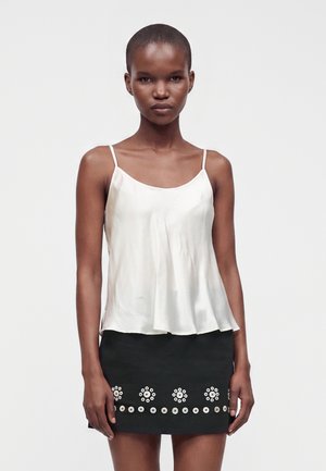 Vrouw met kort haar draagt een witte mouwloze top en een zwarte rok met ronde knoopdecoraties op een effen achtergrond.