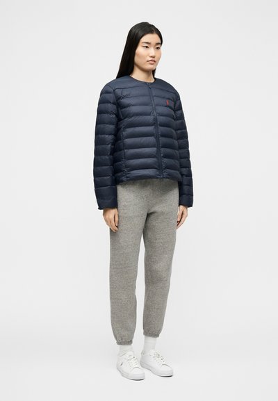 Γυναίκα που στέκεται φορώντας μπλε σκούρο καπιτονέ μπουφάν, γκρι φόρμα, λευκές κάλτσες και λευκά αθλητικά παπούτσια σε απλό φωτεινό φόντο.