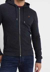 Sweat à capuche zippé bleu marine en tissu doux avec une poche avant, une capuche à cordon de serrage et un petit logo brodé sur la poitrine.