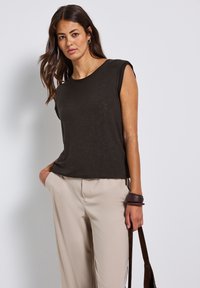Tmavě šedý bezrukávový top s uvolněným střihem, kulatým výstřihem a měkkou strukturou. V kombinaci s volnými béžovými kalhotami a hnědým náramkem.