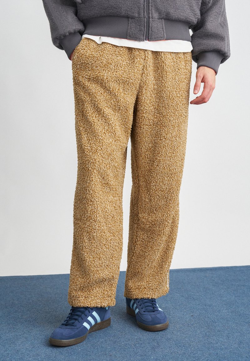 Pantalones de pierna ancha en color beige con textura de forro polar y una superficie suave. Combinados con zapatillas de deporte en color azul marino con rayas en azul claro.