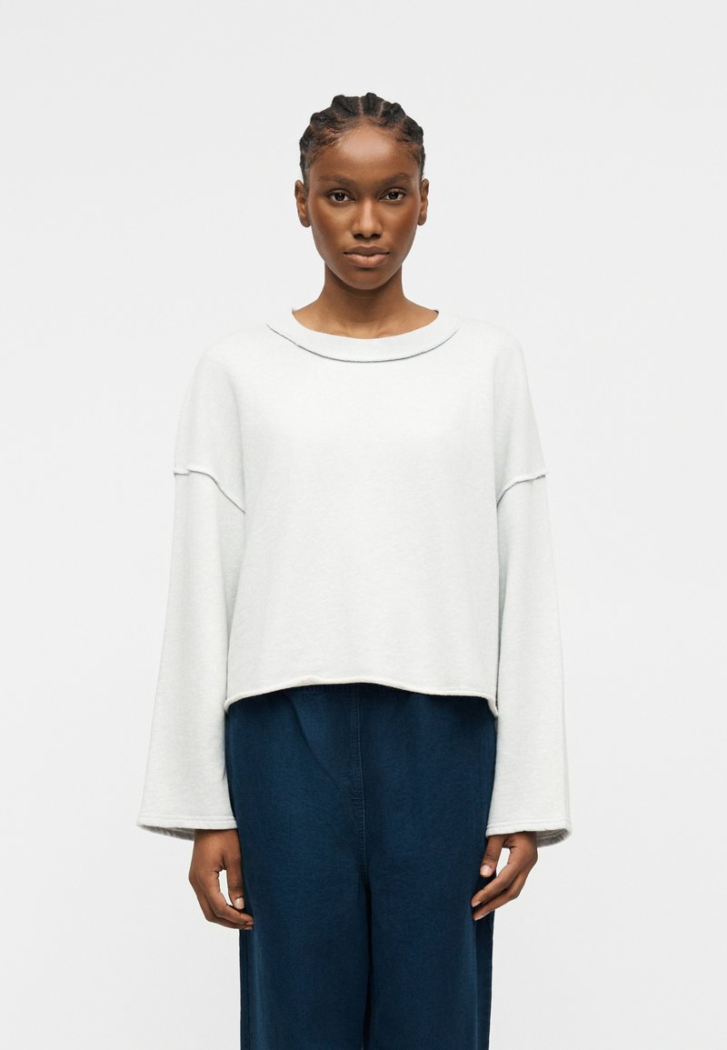 Kobieta z zaplecionymi warkoczami, ubrana w luźną białą bluzkę z długim rękawem i ciemne, szerokie spodnie, stojąca na tle jednolitego jasnego tła.