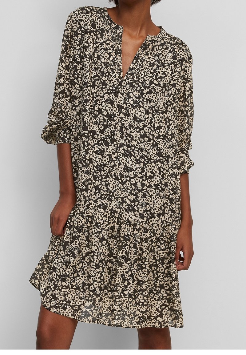 Femme portant une robe noire à fleurs blanches, manches longues et col en V, debout devant un fond uni.
