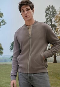 Light brown zip-up sweatshirt with ribbed cuffs and hem, displayed with a neutral-toned undershirt and gray jeans. Smooth texture, casual design.