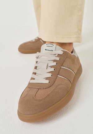 Gros plan sur des baskets Tamaris en daim beige-brun, à lacets blancs et semelles en caoutchouc, portées avec un pantalon crème. Une deuxième chaussure apparaît à l’arrière-plan.