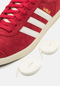 Baskets en daim rouge avec trois bandes blanches, finition texturée et lettrage doré. Comprend deux lacets ronds blancs à côté de la chaussure.