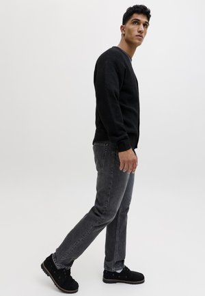 Jeune homme debout de profil, portant un pull noir, un jean gris et des chaussures noires, sur un fond blanc uni.