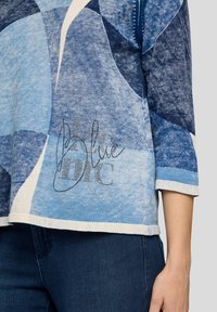 Blauer Pullover mit geometrischem Muster in verschiedenen Blautönen, mit einem weißen Saum und dekorativen silbernen Stickdetails.