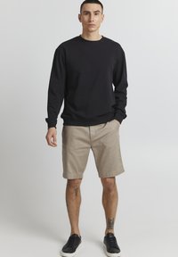 Sort sweatshirt med lange ærmer, parret med beige shorts. Modellen har sorte sneakers på. Enkel, afslappet design med minimale accenter.