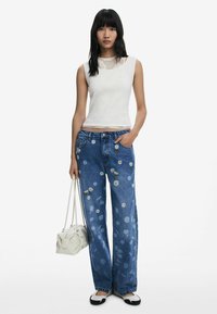 Witte geribde tanktop, blauwe wijde jeans met bloemenborduursel, zwart-witte instapschoenen, met een gestructureerde witte handtas.