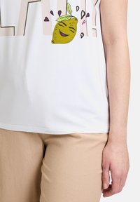 Witte t-shirt met een afbeelding van een glimlachende citroen met groene bladeren en roze accenten, gecombineerd met lichtbeige broeken.