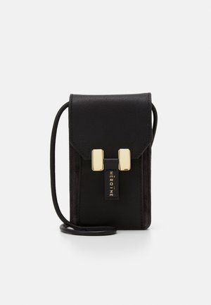 Mała czarna skórzana torba na ramię z prostokątnymi złotymi zapięciami i napisem "HEROINE" na przednim pasku.