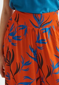 Pantalons fluides orange avec une taille smockée, arborant un motif de feuilles noirs et bleus. Le tissu semble léger et doux.