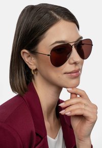 Brūnas aviatora saulesbrilles ar plānu melnu rāmi. Modelis valkā bordoīgu žaketi un zelta aplīšus auskarus, parādot dabiska make-up izskatu.