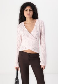 Rosa Spitzentop mit langen Ärmeln und V-Ausschnitt, kombiniert mit braunen, maßgeschneiderten Hosen mit Doppelknopfdetail an der Taille.