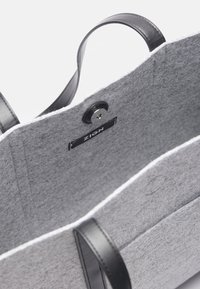 Grå tote-väska i filt med svarta läderhandtag, utrustad med knappstängning och en logomärkt etikett inuti. Slät textur med strukturerad design.