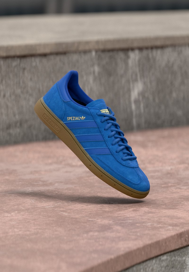 Μπλε αθλητικό παπούτσι Adidas Spezial με σόλα από καουτσούκ, δεμένα κορδόνια, αιωρείται πάνω από λεία πέτρινη επιφάνεια κοντά σε γκρίζο τσιμεντένιο τοίχο.