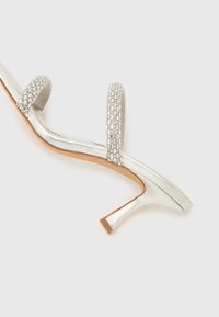 Ασημί πέδιλο με ψηλό τακούνι, δύο λουριά με πολύτιμους λίθους, κυρτό μεταλλικό σχέδιο, μυτερή μύτη και κομψή, υφασμάτινη σόλα.