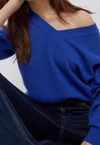 Pull en tricot bleu avec un large col en V, une texture douce et des coutures d'épaule tombantes, assorti à un jean en denim foncé.