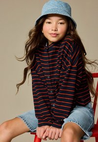 Marineblauw en rood gestreept overhemd met knopen en lange mouwen, gecombineerd met spijkershorts. Het personage draagt een lichtblauwe bucket hat met stippen.