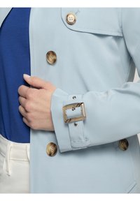 Lichtblauwe jas met grote, glanzende bruine knopen, een mouw met riem en rechthoekige gesp, en een soepele stoftextuur.