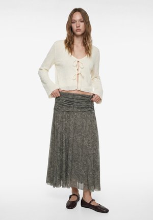 Jonge vrouw staand, gekleed in een crèmekleurige longsleeve top met strik aan de voorkant, een grijze midi-rok met patroon, en donkere Mary Jane schoenen tegen een witte achtergrond.