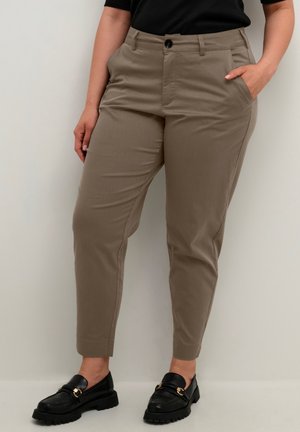 Person iført taupe-farvede ankellange bukser, sorte loafers og en sort top, stående med én hånd i lommen mod en ensfarvet baggrund.