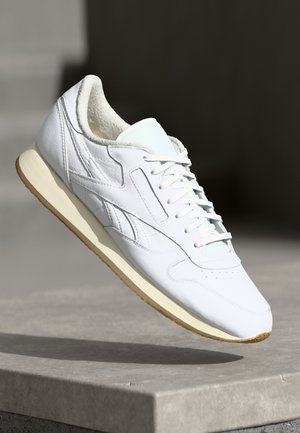 Baskets en cuir blanc avec semelle crème et doublure polaire, montrées flottant au-dessus d'un rebord en béton sur un fond sombre flou.
