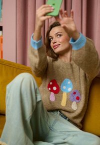 Brązowy sweter wełniany z kolorowymi wzorami grzybów w odcieniach czerwieni, niebieskiego i fioletowego. Jasnoniebieskie mankiety. Widoczne niebieskie dżinsy i zielony telefon.