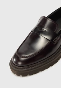 Mørk burgunder lær loafer med en glat finish, som har en dekorativ rem, kontrasterende syninger og en tyk, struktureret gummisål.