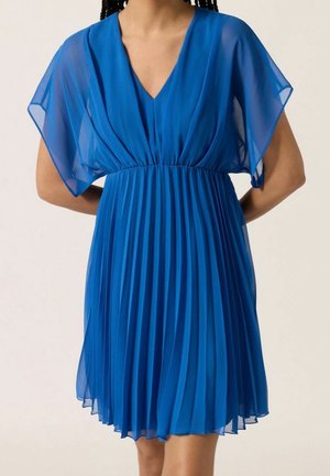 Kobieta w niebieskiej plisowanej sukience z dekoltem w serek i przezroczystymi rękawami w kształcie skrzydeł motyla, stojąca z rękami za plecami.