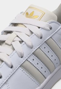 Λευκό αθλητικό παπούτσι Adidas σε κοντινό πλάνο που δείχνει τα κορδόνια, τη υφασμάτινη γλώσσα με χρυσό λογότυπο και τις τρεις λευκές ρίγες με οπές στο πλάι.