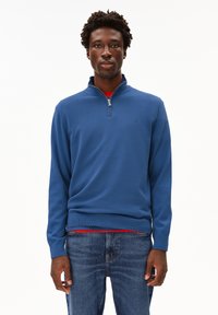 Maglione blu con collo zip realizzato in tessuto a maglia, dotato di polsini e orlo a costine, abbinato a una maglietta rossa, indossato con jeans blu.