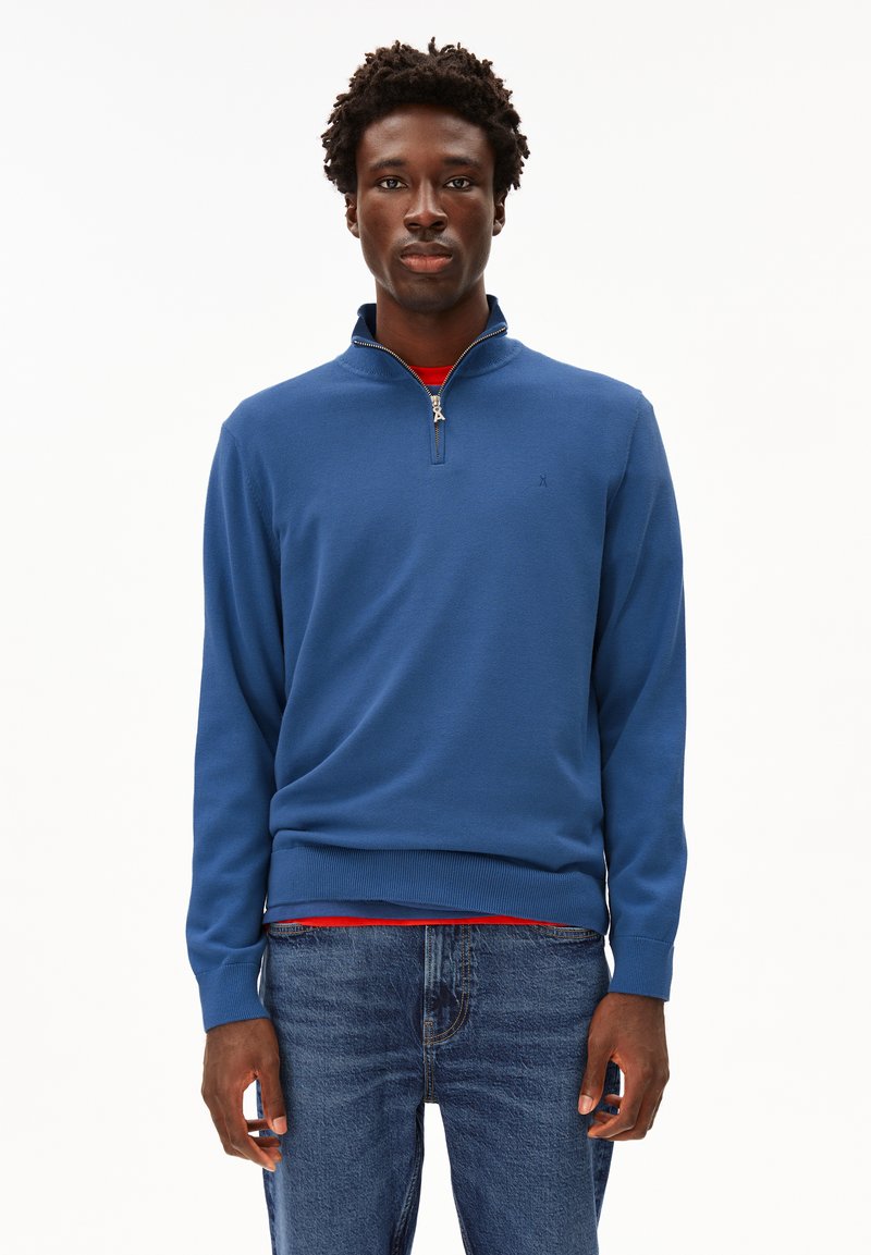 Maglione blu con collo zip realizzato in tessuto a maglia, dotato di polsini e orlo a costine, abbinato a una maglietta rossa, indossato con jeans blu.