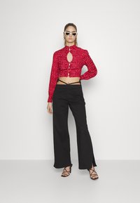 Top cropped vermelho com padrão, apresentando um recorte em forma de chave, combinado com calças pretas de cintura alta com fendas laterais e sandálias de salto pretas com tiras.