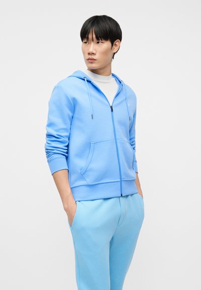 Νεαρός άνδρας που φοράει ανοιχτό μπλε φούτερ με φερμουάρ και αντίστοιχο παντελόνι, στέκεται με τα χέρια στις τσέπες, κοιτώντας προς τα πλάγια σε λευκό φόντο.