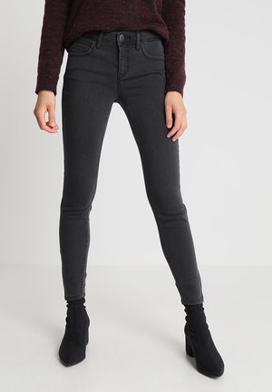 Schwarze Slim-Fit-Jeans kombiniert mit schwarzen Stiefeletten mit Absatz und einem dunkelroten Pullover, von der Taille bis zu den Füßen vor einem schlichten Hintergrund gezeigt.