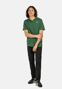 Grünes Polohemd mit Kragen und Logo, kombiniert mit schwarzen Hosen und schwarzen Sneakers mit weißen und tan Akzenten. Eine lässige Passform ist sichtbar.