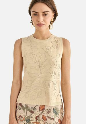 Mujer con top beige sin mangas con patrones abstractos de hojas en relieve y falda con estampado floral, con pendientes de aro dorados.