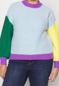 Pull tricoté coloré avec un corps bleu clair, une manche gauche verte, une manche droite jaune et un col et un ourlet côtelés violets.